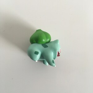 Puede incluir: Una figurita de plástico verde y azul de un personaje Pokemon, Bulbasaur. La figurita está en una pose de carrera con la boca abierta.