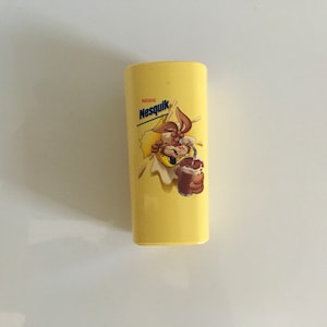 Puede incluir: Un recipiente de plástico amarillo con un conejo de dibujos animados bebiendo chocolate con leche. El recipiente tiene el logotipo de Nestle Nesquik y un plátano en el lateral.