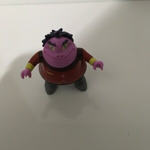 May include: Purple and brown plastic toy figure with a toothy grin and yellow eyes. The figure has a dark purple head with black hair and a brown torso with a yellow belt.