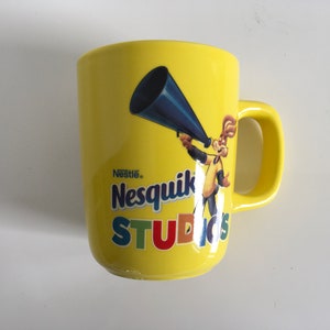 Puede incluir: Taza de cerámica amarilla con un logotipo de Nesquik Studios y un conejo de dibujos animados sosteniendo un megáfono. El logotipo está en colores arcoíris.