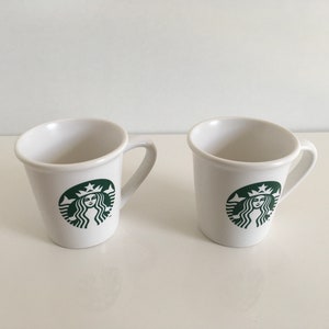 Puede incluir: Dos tazas de cerámica blanca con un logotipo verde de Starbucks que presenta la cara de una mujer.