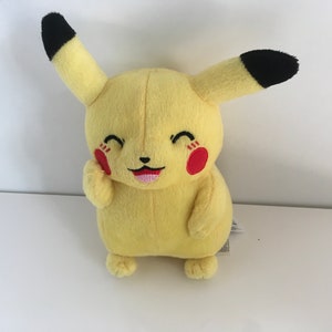 Può includere: Peluche giallo di Pikachu di Pokemon, con orecchie nere e un viso sorridente. Il giocattolo ha le guance rosse e una lingua rosa.