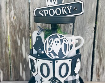 May include: A two-tiered black metal tray decorated with Halloween-themed items including a sign reading SPOOKY St, a circular sign with the word BOO and ghost graphics, a banner spelling BOO, a small ghost mug, and various black and white accents.