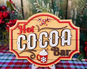 May include: Wooden "Hot Cocoa Bar" sign with red, white, and brown accents. The sign features the words "Hot Cocoa Bar" in layered lettering, candy canes, and a mug with a snowflake design. The sign is on a stand.