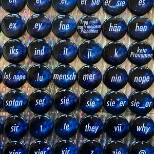 Puede incluir: Una colección de pines redondos y negros con pronombres y un diseño de galaxia azul. Cada pin presenta una palabra o símbolo blanco, como "they", "she", "he" y "xier". Los pines están dispuestos en una cuadrícula.