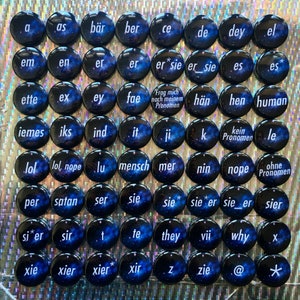 Puede incluir: Un conjunto de 49 botones redondos con fondo negro y diseño de nebulosa azul. Cada botón tiene una palabra diferente impresa en blanco, incluyendo "a", "as", "bär", "ber", "ce", "de", "dey", "el", "em", "en", "er", "er*", "er sie", "er_sie", "es", "es*", "ette", "ex", "ey", "fae", "Frag mich nach meinem Pronomen", "hän", "hen", "human", "iemes", "iks", "ind", "it", "ji", "k", "kein Pronomen", "le", "lol", "lol, nope", "lu", "mensch", "mer", "nin", "nope", "ohne Pronomen", "per", "satan", "ser", "sie", "sie*", "sie er", "sie_er", "sier", "si*er", "sir", "t", "te", "they", "vii", "why", "x", "xie", "xier", "xier*", "xir", "z", "zie", "@", "*".
