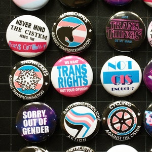 May include: A collection of 20 round buttons with various designs and messages related to transgender identity and rights. The buttons feature a variety of colors, including pink, blue, white, black, and rainbow. Some of the messages on the buttons include "We Want Trans Rights", "Not Your Opinion", "Just Trans Things", "Not Cis Enough", "Snowflakes Against the Cistem", and "Attack Helicopters Against Trans Discrimination".