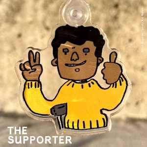 Könnte beinhalten: Eine gelbe Comicfigur mit braunen Haaren und Haut, die einen gelben Pullover mit einer schwarzen Tasche trägt, zeigt mit einer Hand ein Peace-Zeichen und mit der anderen einen Daumen nach oben. Der Text "THE SUPPORTER" befindet sich unter der Figur.