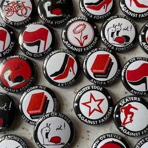Peut inclure: Une collection de boutons ronds noirs et blancs avec des graphismes rouges et blancs. Les boutons présentent des slogans tels que "Antifa Forever", "Now or Never", "Against Fascism", "Vegan", "Queer Edge", "Skater", et "Action".