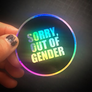Può includere: Un adesivo olografico rotondo con la scritta "SORRY, OUT OF GENDER" in bianco e un gradiente arcobaleno. L'adesivo ha uno sfondo nero e un bordo arcobaleno.