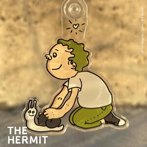 Könnte beinhalten: Eine Comicfigur mit grünem Haar kniet und hält eine weiße Schnecke mit einem lächelnden Gesicht. Die Figur trägt ein weißes Hemd und eine grüne Hose. Der Text "THE HERMIT" befindet sich am unteren Rand des Bildes.