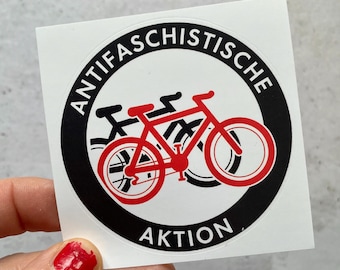 May include: A black and white sticker with the text "Antifachistische Aktion" in a circle around a red silhouette of three bicycles.