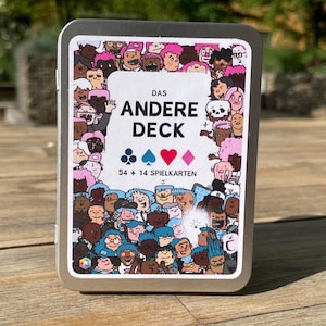 Op de afbeelding: Een rechthoekig zilverkleurig metalen blik met een kaartspel. Het etiket bevat een kleurrijke illustratie van diverse stripfiguren en de tekst "DAS ANDERE DECK" met de symbolen voor klaveren, schoppen, harten en ruiten.