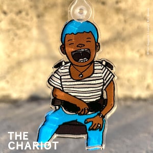 Könnte beinhalten: Ein blau-weiß gestreiftes Hemd mit einem blauen Stirnband und einer blauen Jeans. Die Person sitzt auf einem Stuhl und hat ein breites Lächeln im Gesicht. Der Text "THE CHARIOT" befindet sich am unteren Rand des Bildes.