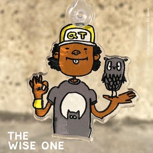 Könnte beinhalten: Eine Comicfigur trägt einen gelben Hut mit den Buchstaben "QT" darauf, ein graues Shirt mit einem weißen Kreis und einer schwarzen Katze im Kreis und eine graue Eule auf dem Arm. Der Text "THE WISE ONE" befindet sich unter der Figur.