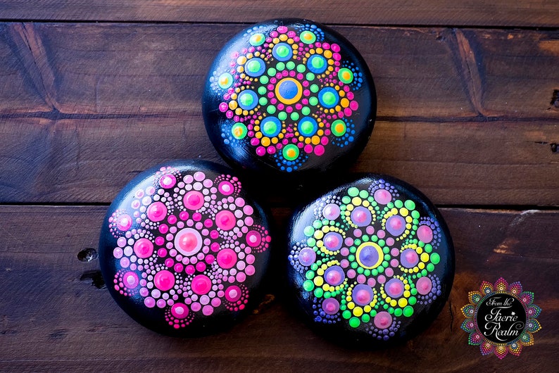May include: Three round black stones painted with colorful mandala designs. The designs feature bright pink, green, blue, yellow, and purple dots.