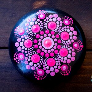 May include: A black stone painted with a pink and white mandala design. The mandala is a circular pattern with a central circle and radiating lines of dots.