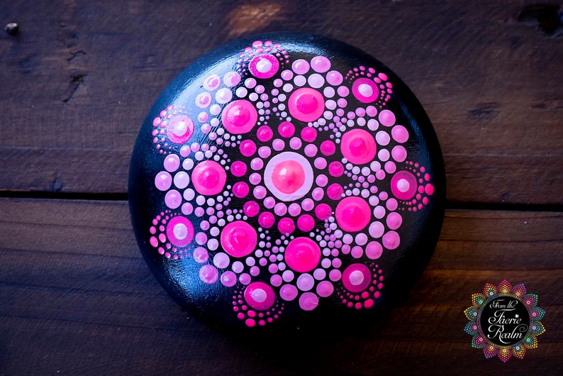 May include: A black stone painted with a pink and white mandala design. The mandala is a circular pattern with a central circle and radiating lines of dots.