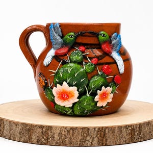 May include: A brown ceramic mug featuring a raised, hand-painted design of two hummingbirds facing each other above a green cactus with white spines, orange flowers, and a small orange butterfly on the side, complete with a curved handle.