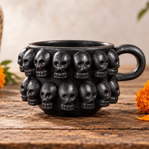 May include: A black ceramic mug featuring a textured surface adorned with two horizontal rows of sculpted human skulls encircling the exterior. The mug includes a single curved handle on the right side and has a matte finish throughout the design.