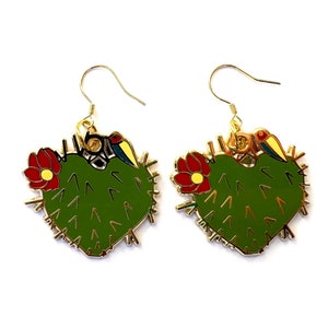 May include: A pair of gold-tone earrings featuring a green cactus design with red flowers and a small bird. The earrings have a hook closure and are set against a white background. The cactus shape is approximately 1.5 inches wide.
