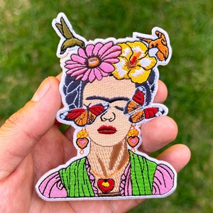May include: Embroidered iron-on patch featuring a colourful portrait of Frida Kahlo with butterflies over her eyes, flowers in her hair, and a heart-shaped pendant.