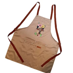May include: A tan canvas apron with brown leather straps and a pocket. The apron features a colourful illustration of Frida Kahlo with flowers in her hair and butterflies on her face.