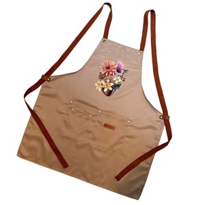 May include: A tan canvas apron with brown leather straps and a floral print on the front. The apron has a large pocket on the front with a small pocket above it.