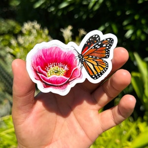 May include: A sticker featuring a pink flower with a monarch butterfly landing on it. The sticker is on a hand against a green background.