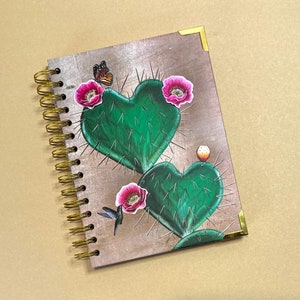 May include: A spiral bound notebook with a brown cover featuring a green cactus with pink flowers and a butterfly. The cactus is shaped like a heart.