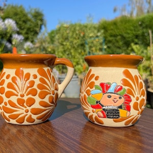 May include: Two ceramic mugs with a rustic design. One mug has a handle and is decorated with orange floral patterns. The other mug features a colorful doll design. Both mugs have a brown rim and base.