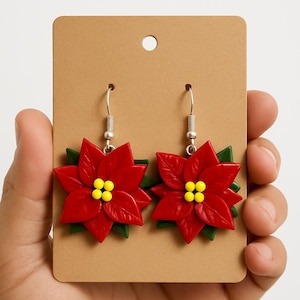 May include: A pair of poinsettia earrings. Each earring features a vibrant red flower with green leaves and yellow center accents. The earrings are displayed on a brown card with a small hole at the top.