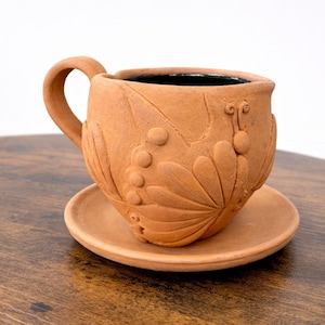 May include: A terracotta-colored ceramic teacup and saucer set. The cup features a handle and is adorned with embossed floral and berry designs. The set is placed on a wooden surface, showcasing the handcrafted details of the pottery.