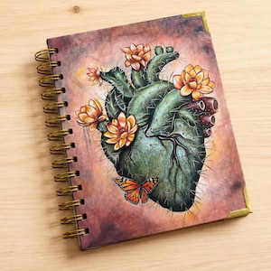 Puede incluir: Un cuaderno de espiral con una ilustración en la portada de un corazón anatómico con forma de cactus, decorado con flores naranjas y una mariposa monarca. El cuaderno tiene esquinas metálicas doradas y una espiral de alambre a juego.