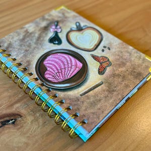 May include: A brown spiral bound journal with a gold coil. The cover features a pink heart-shaped design on a plate, a butterfly, and a heart-shaped design with a white center.