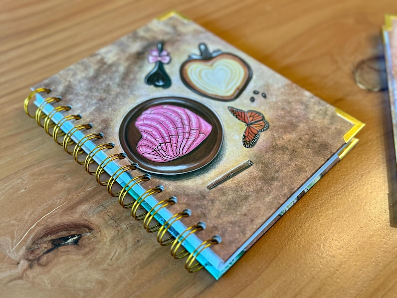 May include: A brown spiral bound journal with a gold coil. The cover features a pink heart-shaped design on a plate, a butterfly, and a heart-shaped design with a white center.