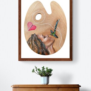 May include: A painting on a wooden artist's palette depicting a woman with long brown hair looking up at a hummingbird. The hummingbird is flying towards the woman with a red paper heart attached to its tail by a string.