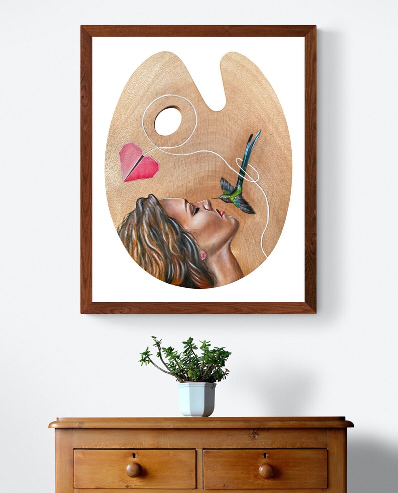 May include: A painting on a wooden artist's palette depicting a woman with long brown hair looking up at a hummingbird. The hummingbird is flying towards the woman with a red paper heart attached to its tail by a string.