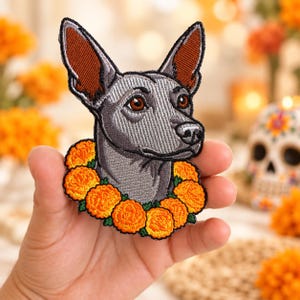 Puede incluir: Un parche bordado que muestra la cabeza de un perro gris con orejas grandes y erguidas y ojos marrones. El perro está rodeado por una corona de flores de cempasúchil de color naranja vibrante con pequeñas hojas verdes, en hilo detallado.
