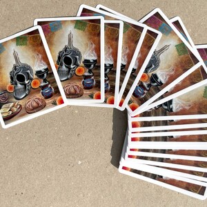 May include: A set of 15 stickers featuring a black and white skull with a candle on top, surrounded by orange fruit, a small cup of coffee, and a traditional Mexican bread. The background is a brown wall with colorful paper streamers.
