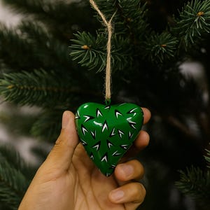 May include: A green heart-shaped Christmas ornament with white and black accents. The ornament is suspended by a natural twine string and is set against a backdrop of evergreen branches. The heart is approximately 3 inches wide.