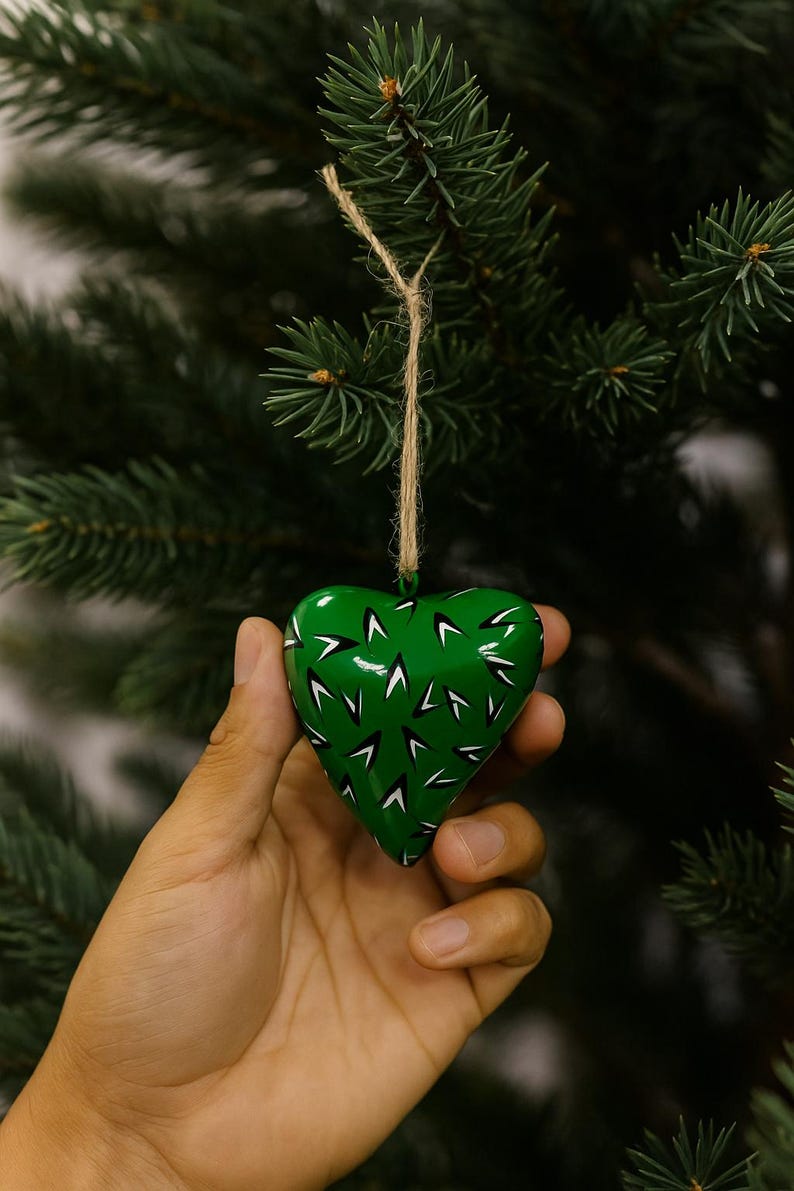 May include: A green heart-shaped Christmas ornament with white and black accents. The ornament is suspended by a natural twine string and is set against a backdrop of evergreen branches. The heart is approximately 3 inches wide.