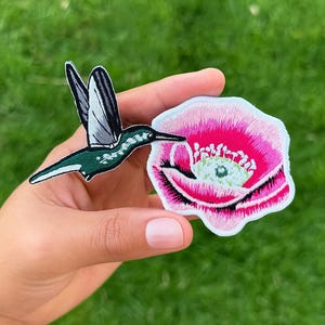 May include: Embroidered patches featuring a hummingbird and a flower. The hummingbird is green, black, and white, with detailed wing patterns. The flower is a vibrant pink with a white and green center. The patches are on a white background.