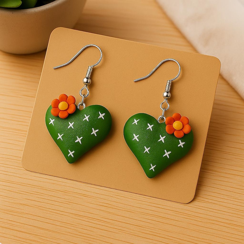 May include: A pair of green heart-shaped earrings with white cross details, resembling a cactus. Each earring features a small orange flower with a yellow center. The earrings hang from silver-colored hooks.