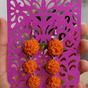 Puede incluir: Pendientes de cal&eacute;ndula naranja con ganchos plateados, exhibidos en un recorte de papel morado. Cada pendiente presenta tres esferas naranjas texturizadas y peque&ntilde;as hojas verdes. El papel morado tiene un dise&ntilde;o floral decorativo.