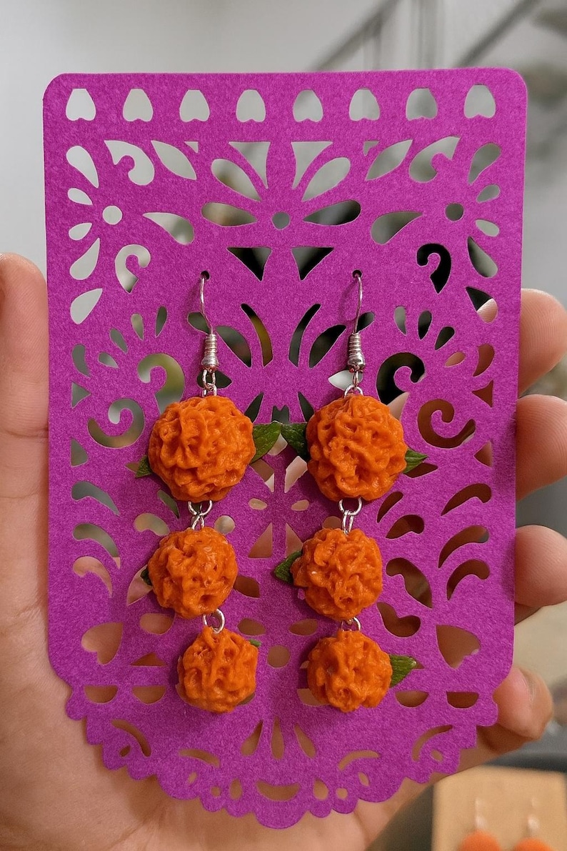 Puede incluir: Pendientes de cal&eacute;ndula naranja con ganchos plateados, exhibidos en un recorte de papel morado. Cada pendiente presenta tres esferas naranjas texturizadas y peque&ntilde;as hojas verdes. El papel morado tiene un dise&ntilde;o floral decorativo.
