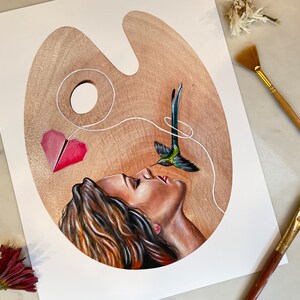 May include: A painting on a wooden palette depicting a woman with her eyes closed, a hummingbird with a string attached to its beak, and a red paper heart. The string connects the hummingbird to the heart and the heart to the woman's nose.