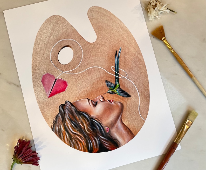 May include: A painting on a wooden palette depicting a woman with her eyes closed, a hummingbird with a string attached to its beak, and a red paper heart. The string connects the hummingbird to the heart and the heart to the woman's nose.