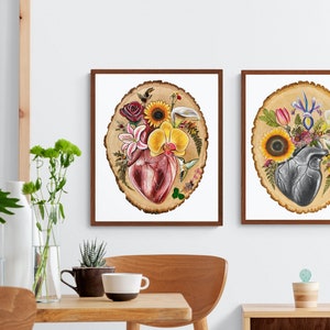May include: Two framed prints of anatomical hearts surrounded by flowers and greenery on a wood slice background. The heart on the left is red and the heart on the right is gray.