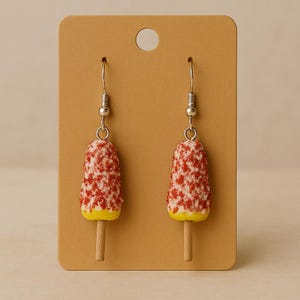 May include: Novelty earrings in the shape of red and white ice lollies with yellow bases and wooden sticks. The earrings are suspended from silver-coloured fish hook ear wires and presented on a brown earring card.